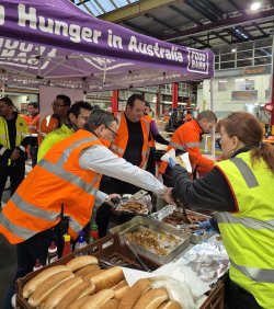 This image captures a lively food distribution event under a marquee at Perth markets, featuring a banner that reads "Hunger in Australia - Foodbank." Volunteers in high-visibility vests are actively serving a variety of hot dishes, including sausages, from metal trays. Fresh bread rolls are also available, highlighting the community's effort to combat hunger. The setting, marked by organization and cooperation, underscores the charitable initiative to feed those in need.