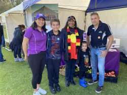 The image captures a group of five people at an outdoor event, standing in front of a tent with a community or cultural theme. From left to right, there’s a woman in a purple shirt, a boy in a colorful hoodie, a woman in an Aboriginal flag scarf with a young girl, and a man in a black and white polo shirt.