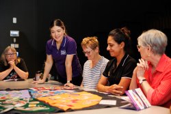 This image captures a group of women engaged in an interactive healthy eating activity. In the centre, a woman wearing a purple Foodbank shirt is leading the session, focusing on colorful materials spread out on the table. The other women, displaying a mix of expressions and engagement, are attentive to the session. They are seated around a black table in what appears to be a modern, dimly lit room, suggesting an informal, yet organized setting for learning or community engagement.