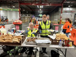 The image depicts a vibrant community event or gathering, likely held in a warehouse or similar indoor setting, as indicated by the industrial environment and signage like "Brian Fresh Produce." Four adults are actively engaged in serving food, dressed in high-visibility safety vests, which suggests a well-organized event, possibly a community service or volunteer effort. In the foreground, a man is holding a hot dog with a smile, surrounded by trays of more hot dogs and sausages, all ready to be served. The table is well-stocked with fresh buns and condiments like ketchup and mustard, catering to the attendees’ tastes. Other participants in the background are busy with various tasks, contributing to the smooth operation of the event.