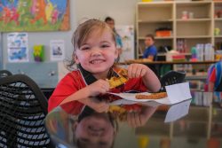 This photo captures a joyful young girl, dressed in a red shirt, sitting at a table in a classroom setting. She is happily eating a slice of toast, holding it with both hands and smiling broadly at the camera. In the background, other children and classroom supplies are visible, along with colorful artwork on the wall, creating a lively and cheerful atmosphere. The reflection of the girl on the shiny table adds an artistic touch to the image.