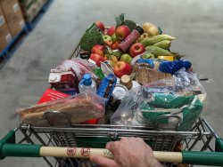 The image shows a shopping trolley filled with a variety of groceries from a first-person perspective, as seen by someone pushing the cart. The cart is packed with an assortment of items including fresh fruits like apples, vegetables such as broccoli and corn, as well as packaged goods like bread, milk cartons, and bottled water.