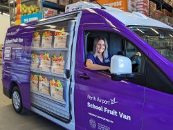 The image shows a smiling woman in the driver's seat of a vibrant purple van, which is labeled "Perth Airport School Fruit Van" and marked with the Foodbank logo. The van is situated inside a warehouse, with shelves of goods visible in the background. The side of the van features a realistic decal depicting crates of fresh fruits, emphasizing its role in distributing healthy food to schools. The woman, wearing a blue Foodbank uniform, looks cheerful and proud, highlighting the positive community impact of this initiative. 