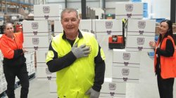 The image shows three people in a warehouse setting. A man in the foreground, wearing a high-visibility vest and gloves, is posing with one hand on his chest. Behind him, two women in high-visibility jackets are smiling and standing near stacks of boxes.