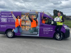 The image features a group of Foodbank employees and volunteers, exuberant and full of energy, gathered around a purple van labeled "Perth Airport School Fruit Van." The van, used for distributing fresh fruit to schools, helps emphasize the community-driven initiative supported by the Perth Airport and Foodbank. The volunteers, clad in orange high-visibility vests, are seen celebrating their efforts—two are joyfully jumping in the air beside the van, another smiles from the driver’s seat, and one proudly displays a box marked "Food for Good." Their expressions and actions convey a strong sense of commitment and enthusiasm for their cause, reflecting the positive impact of their work on school nutrition programs.