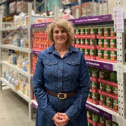 The image features a middle-aged woman standing in front of a grocery store aisle. She is smiling and dressed in a blue denim jacket over a similar blue denim shirt, accessorized with a large brown belt. 