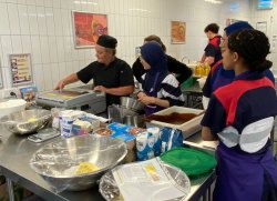 The image shows a busy kitchen setting, likely in a school or community center. It features three individuals: a woman in a black chef's uniform operating kitchen equipment, a young woman in a hijab assisting her, and another young person in a blue and red uniform, looking on. The kitchen is equipped with stainless steel counters and various cooking supplies. The overall scene suggests a hands-on cooking class or food preparation session.